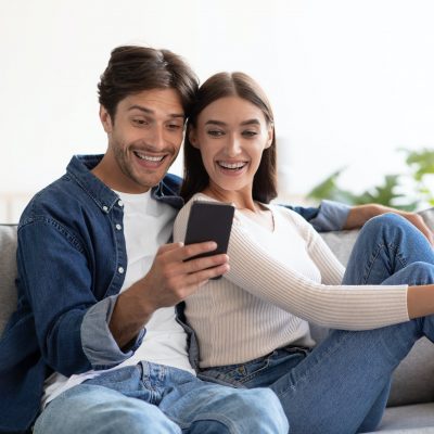Smiling shocked young caucasian husband and wife look at smartphone, have meeting in living room