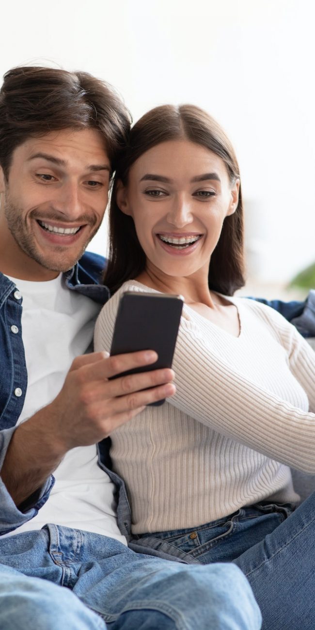 Smiling shocked young caucasian husband and wife look at smartphone, have meeting in living room